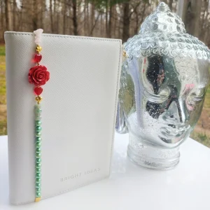 Apple of My Eye bookmark from Inanna Joy Boutique, featuring a red rose charm with green beads, displayed beside a silver Buddha head and white journal.