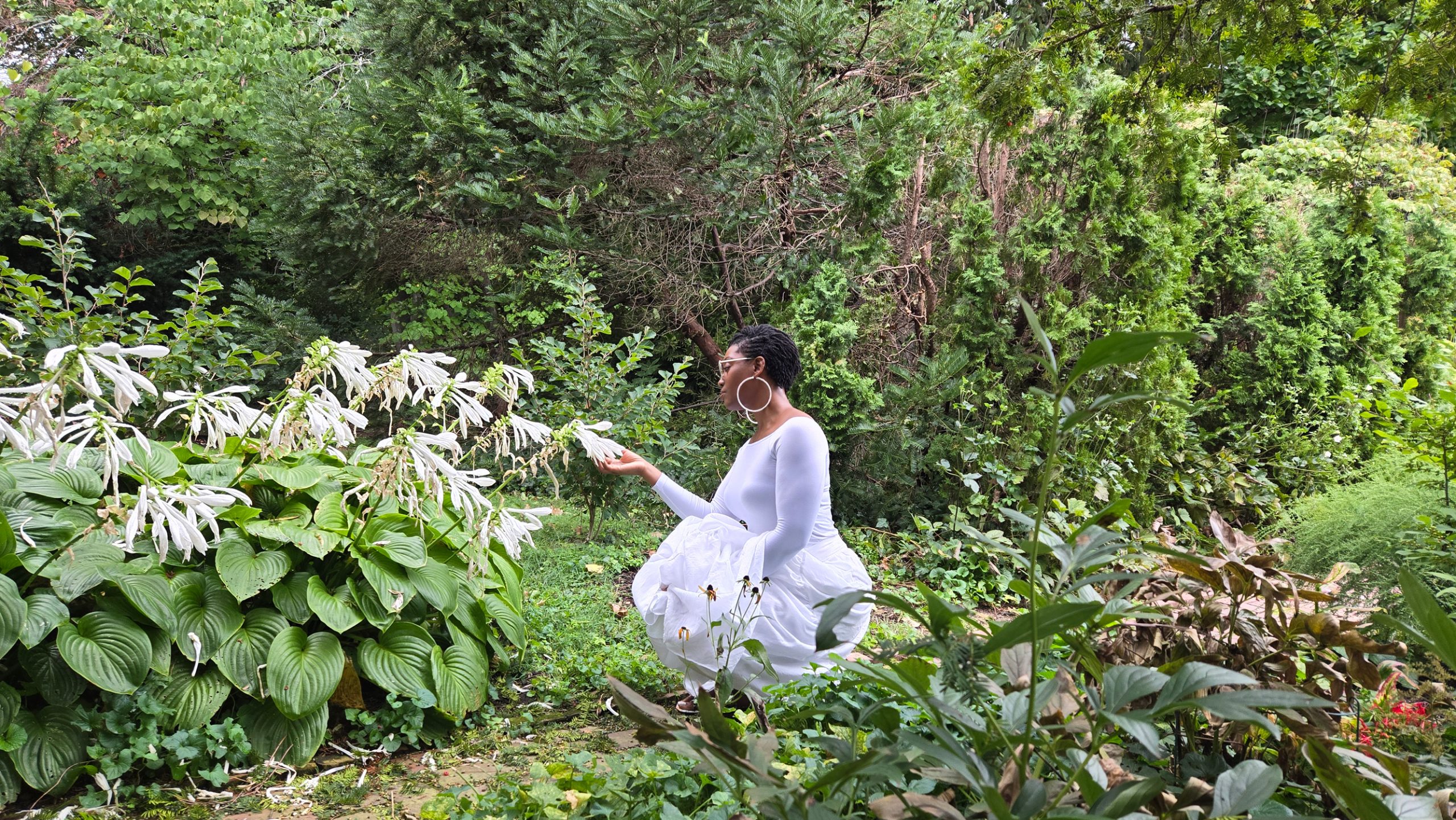 Inanna Joy, motivational author and speaker, sitting in a lush green garden wearing a flowing white dress, reaching toward blooming white flowers.